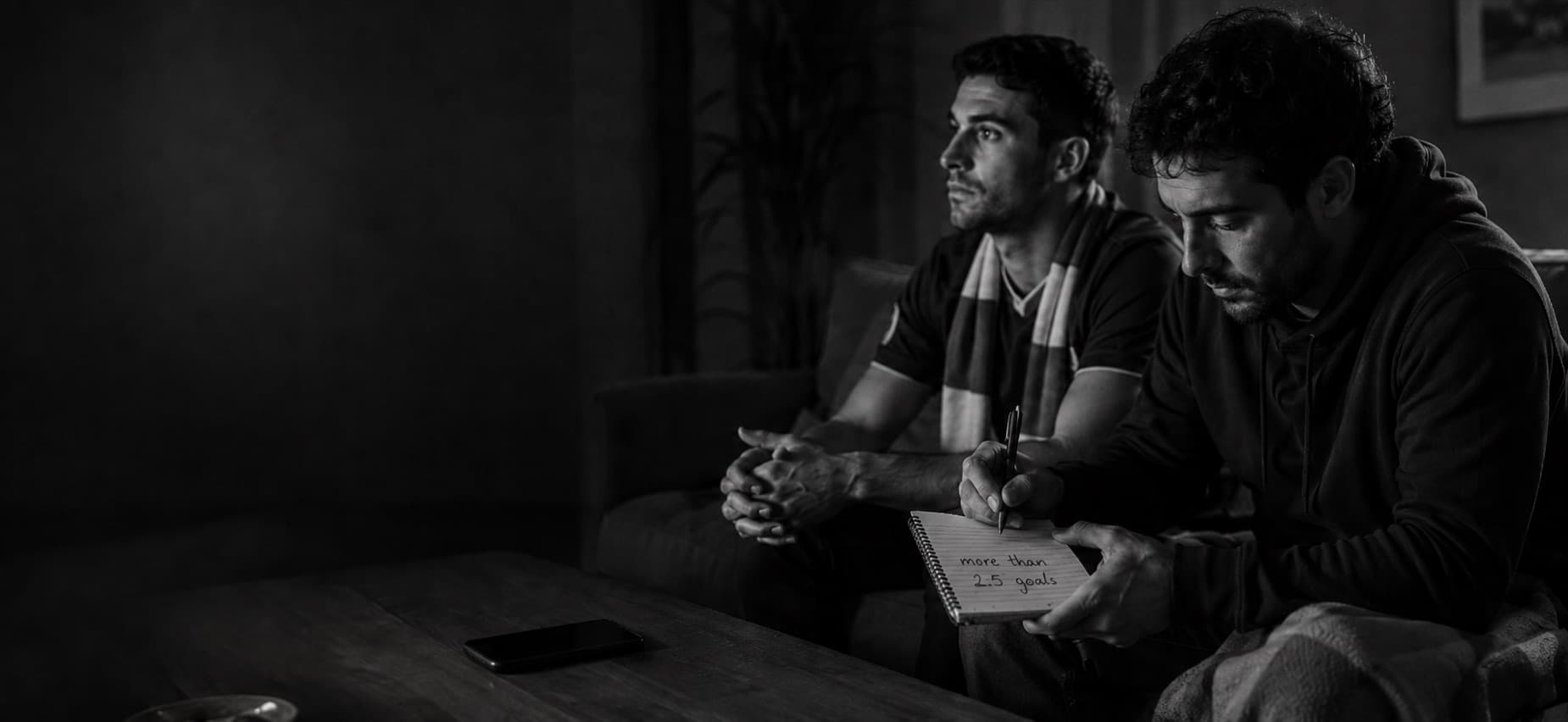 Two fans on a sofa; one writes more than 2.5 goals in a notebook while watching the match